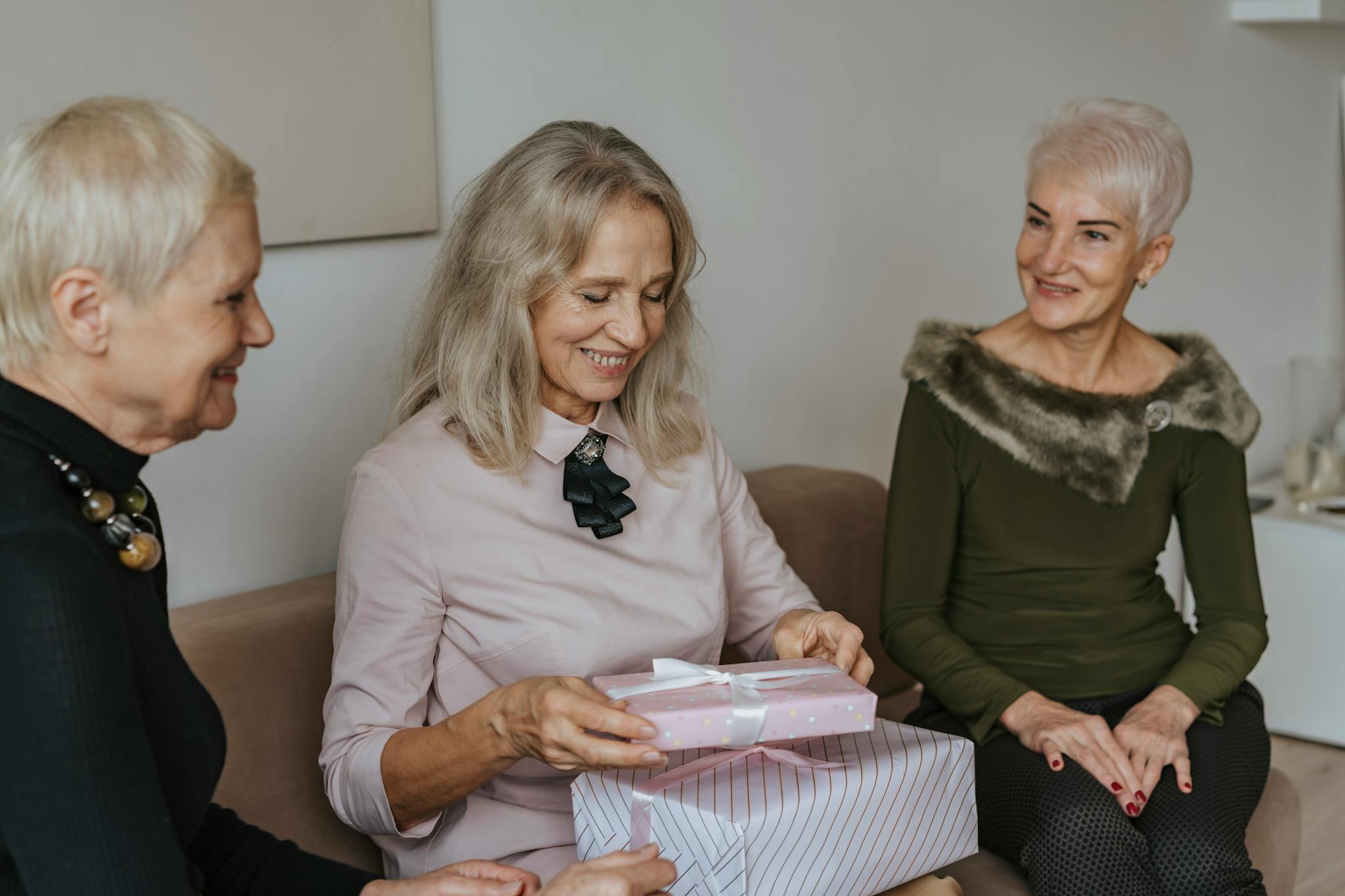 Three elderly women joyfully exchange gifts while sitting indoors, celebrating together.