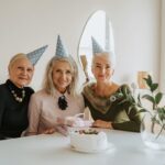 Three elderly women wearing party hats celebrating a birthday with cake and gifts.