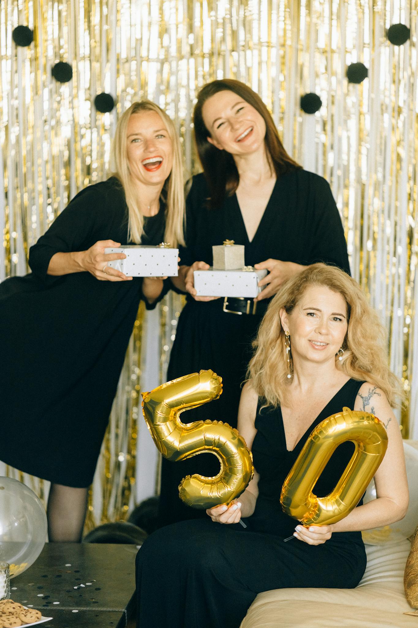 Three women celebrating a 50th birthday with gifts and golden balloons indoors.